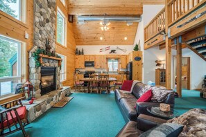 Un intérieur de chalet chaleureux avec une cheminée en pierre, un coin repas en bois et un espace en mezzanine où sont rangés les équipements de ski.