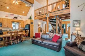 A spacious living area with a high ceiling, wooden beams, a dining table with chairs, a leather sofa, and a staircase leading to an upper level.