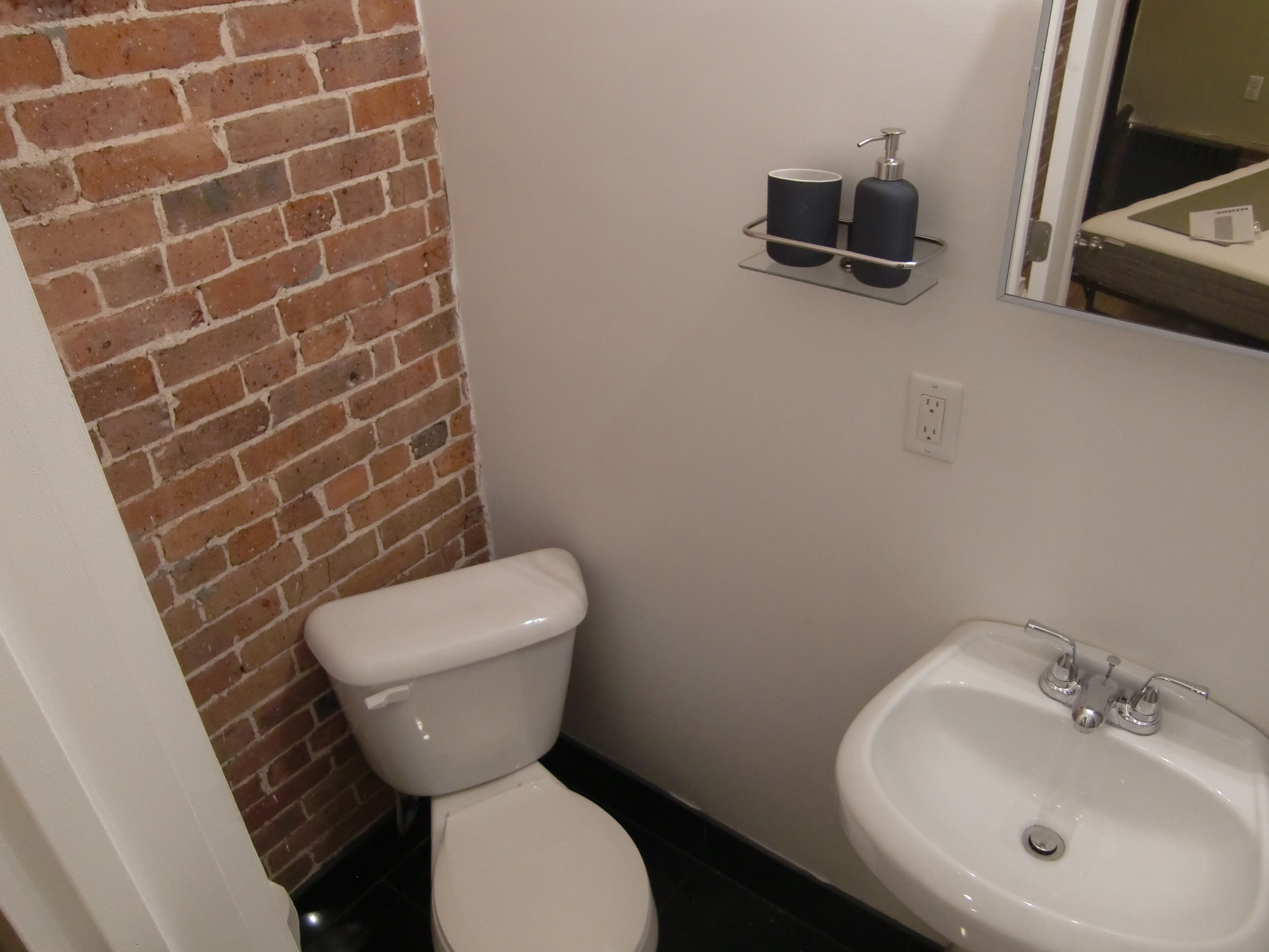 A bathroom with a white toilet, a sink, and a mirror. There is a brick wall and a shelf with toiletries.