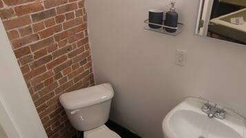 A bathroom with a white toilet, a sink, and a mirror. There is a brick wall and a shelf with toiletries.