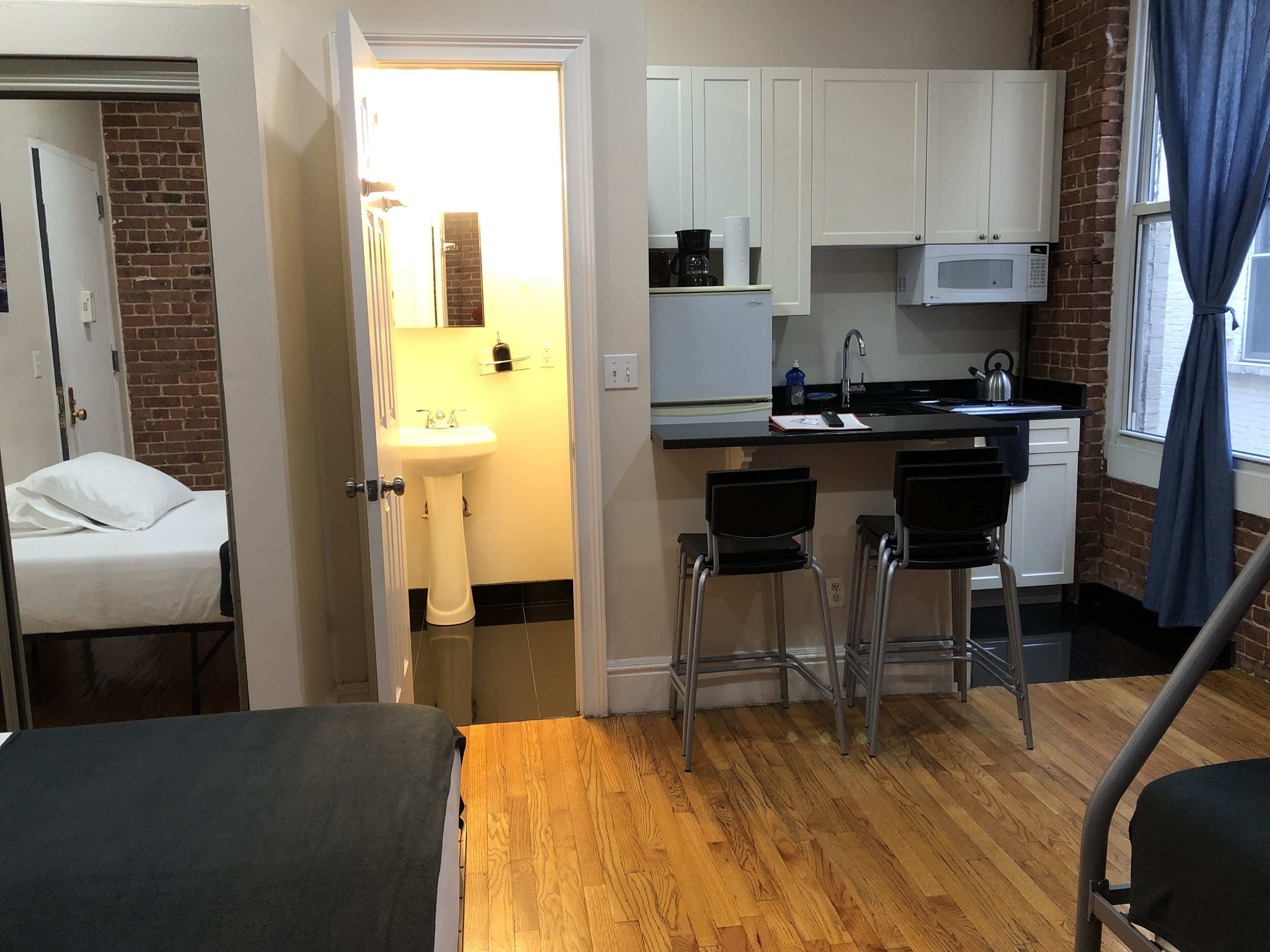 A compact hotel room with a wooden floor, a bed, a small kitchenette with a microwave, a sink, and a window with curtains.