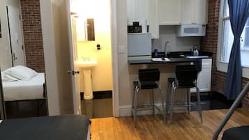 A compact hotel room with a wooden floor, a bed, a small kitchenette with a microwave, a sink, and a window with curtains.