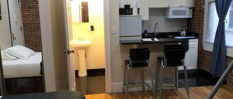 A compact hotel room with a wooden floor, a bed, a small kitchenette with a microwave, a sink, and a window with curtains.