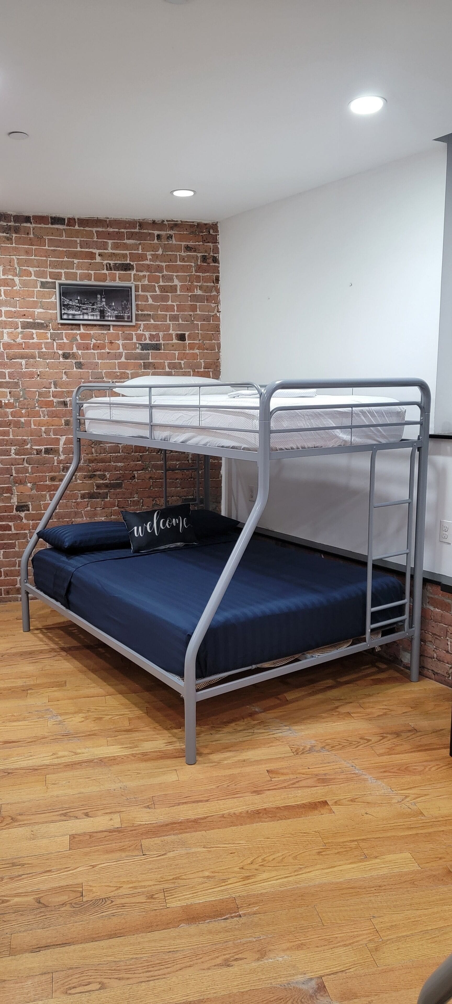 Ein Etagenbett mit Metallgestell und blauer Bettwäsche, eine Ziegelwand mit einem gerahmten Bild und ein Holzboden.