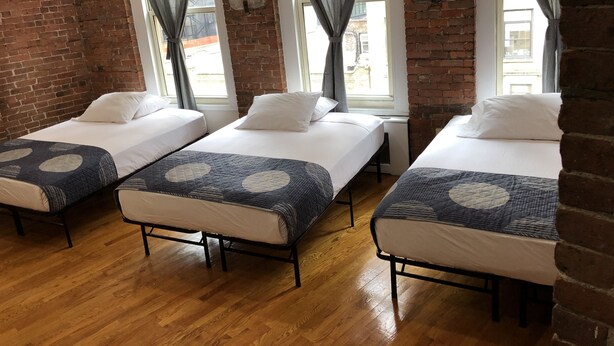 A room with three beds, each with a patterned bedspread and white pillows, set against a brick wall with large windows and sheer curtains. - Studios Midtown Manhattan (New York)