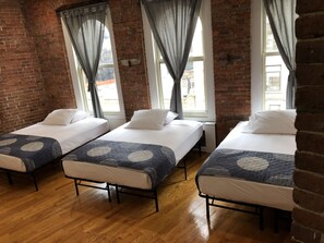 A room with three beds, each with a patterned bedspread and white pillows, set against a brick wall with large windows and sheer curtains. - Studios Midtown Manhattan (New York)