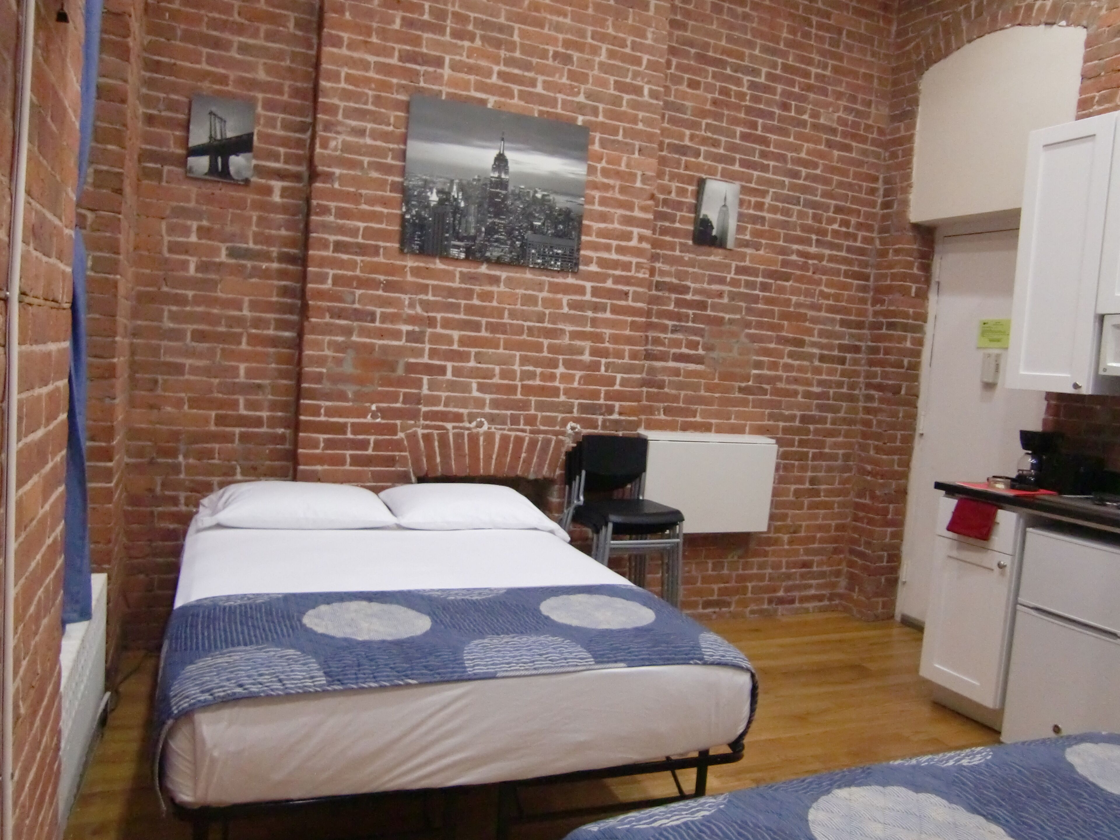 A small, cozy room with a brick wall, a bed with a patterned bedspread, a chair, a desk with a computer, and a small kitchen area.