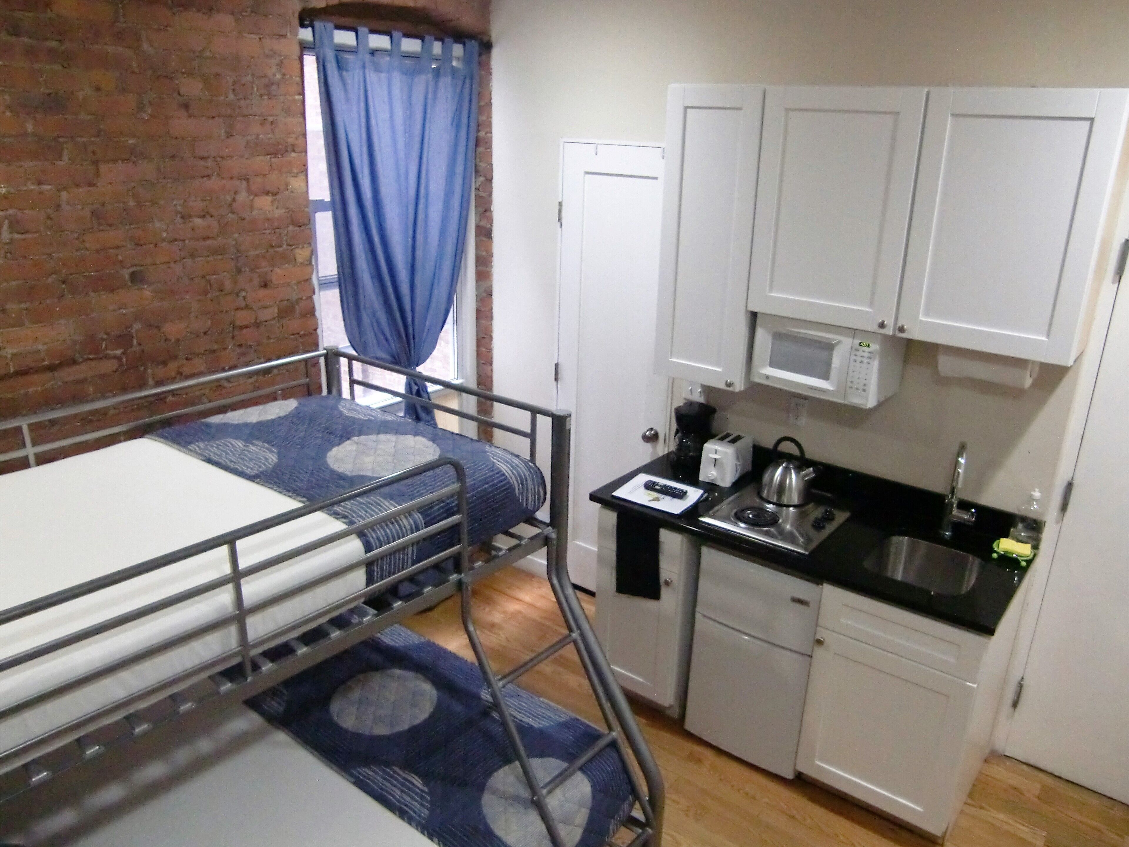 Una habitación pequeña con literas, kitchenette con microondas y un ventanal con cortinas azules.