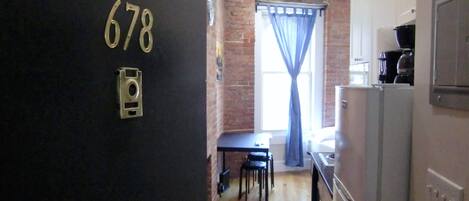 Un pasillo estrecho con una puerta marcada “678” que da a una zona de cocina con refrigerador, cafetera y ventana con cortinas azules.