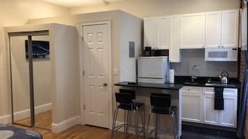 Una cocina compacta con armarios blancos, un refrigerador, un microondas y un lavamanos. Hay dos taburetes de bar y una puerta que da a otra habitación.