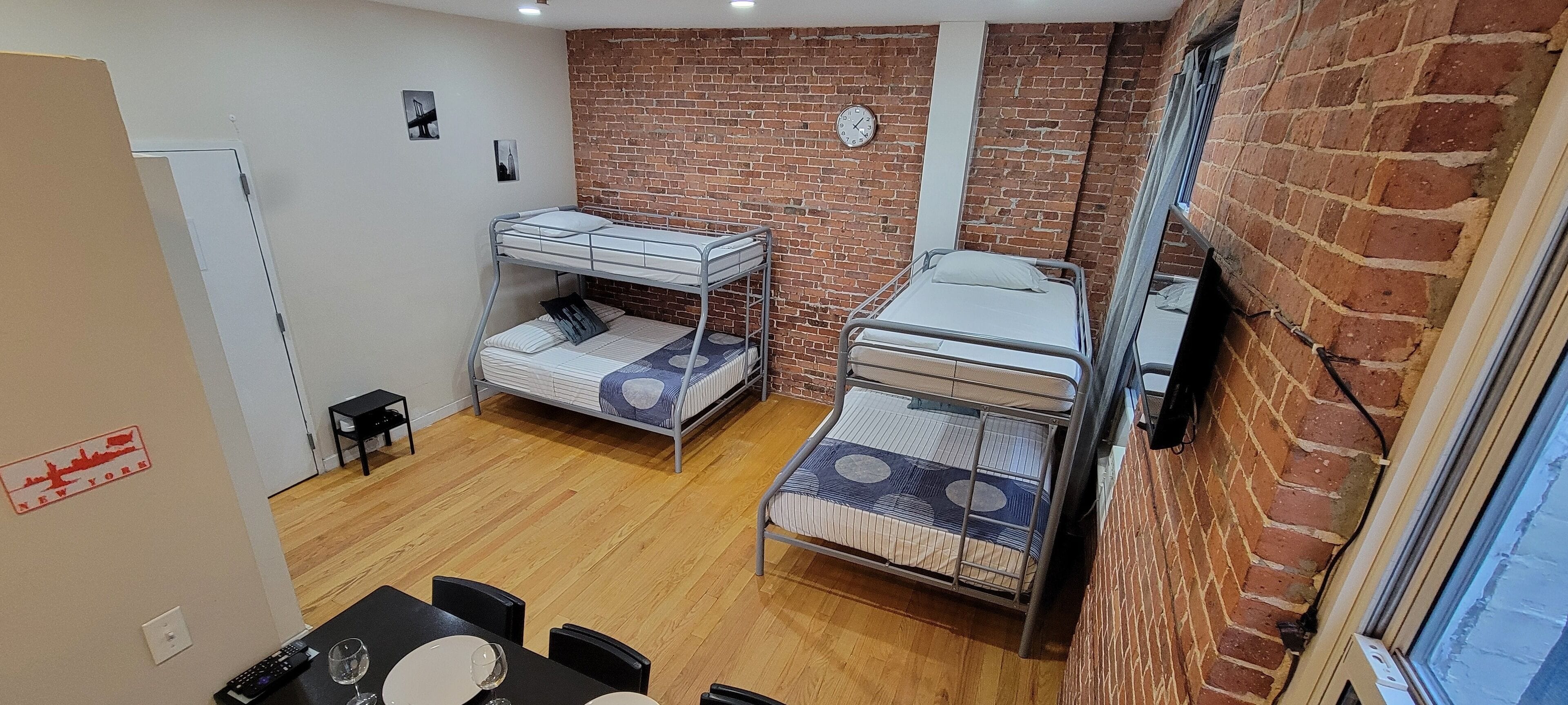 A dormitory room with bunk beds, a brick wall, a clock, and a window.