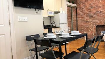 A dining area with a black table set for six, surrounded by black chairs. A flat-screen TV is mounted on the wall above the table.