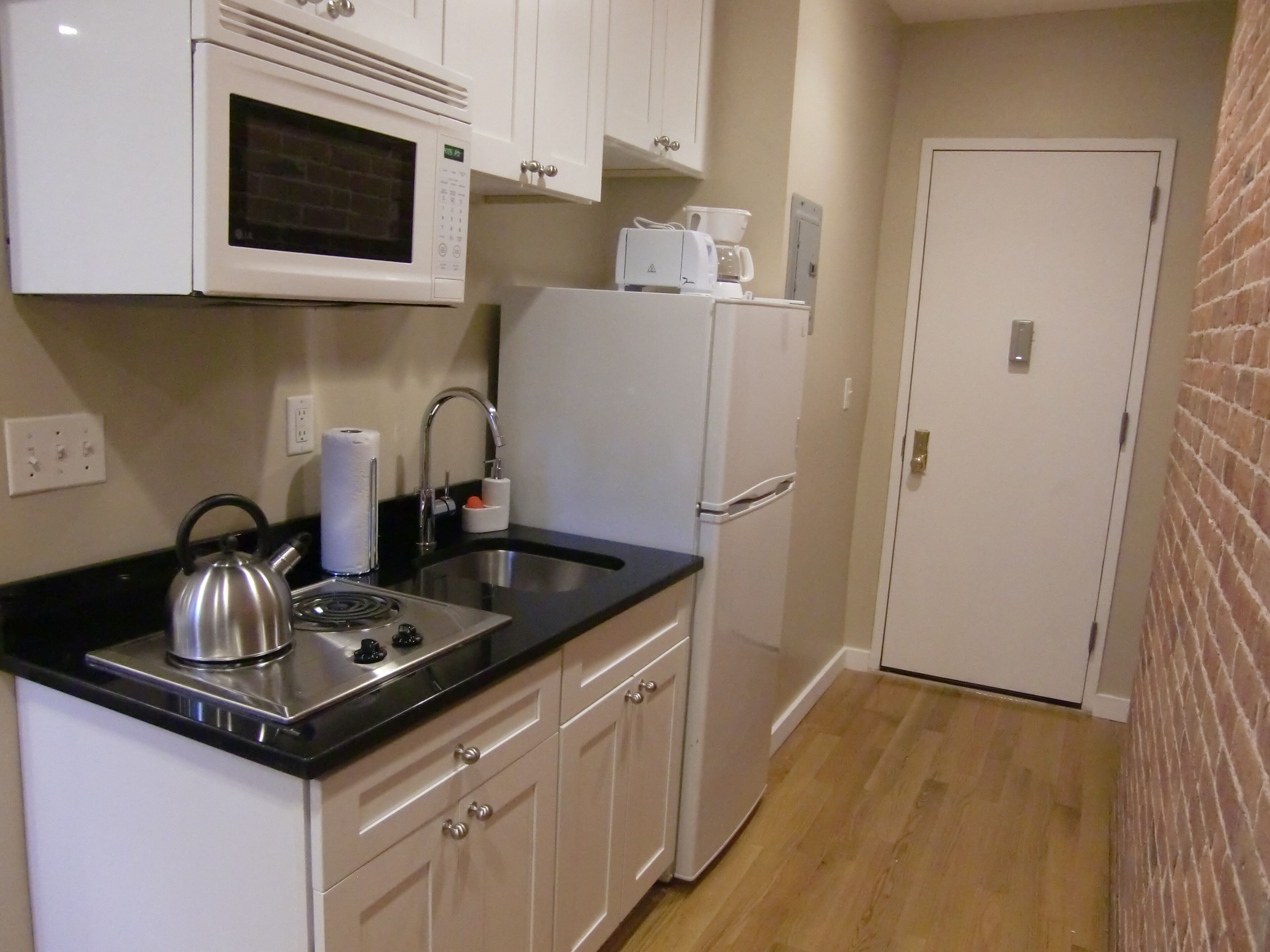 A compact kitchen with white cabinets, a stainless steel kettle, a microwave, a sink, and a refrigerator.