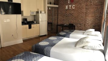 Ein Hotelzimmer mit Küchenzeile, zwei Betten mit blau-weißer Bettwäsche, einer Ziegelwand mit der Aufschrift „I ♥ NYC“ und einem kleinen Tisch mit einer Pflanze.