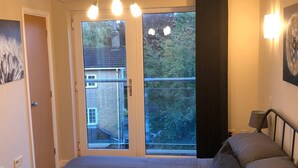 A bedroom with a large bed, a nightstand, a wall-mounted lamp, and a view of a balcony with a door leading outside. - River/Meadow View Apartment - Little Paxton (Little Paxton)