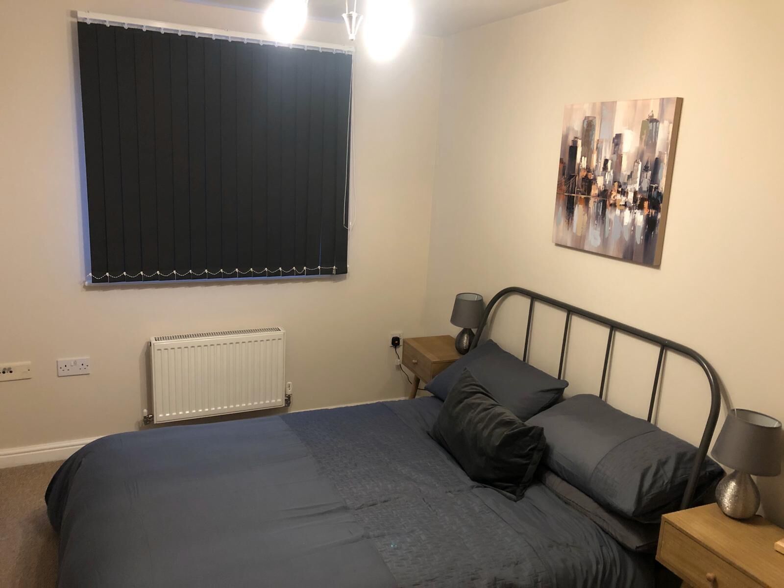 A bedroom with a bed, a nightstand, a lamp, a radiator, a window with curtains, and a painting on the wall.
