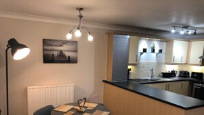 A modern kitchen with a central island, a dining table with chairs, a radiator, and a painting on the wall. - River/Meadow View Apartment - Little Paxton (Little Paxton)