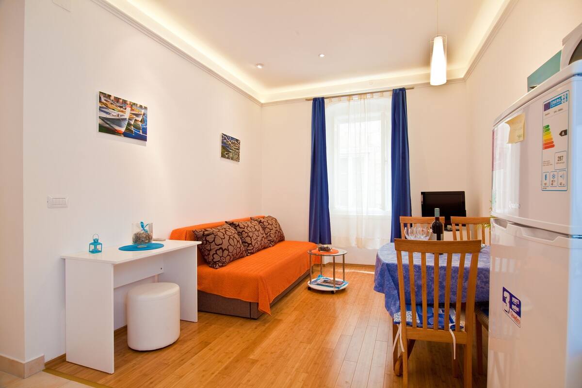A compact living space with a sofa, a small desk, a dining table, and a refrigerator.