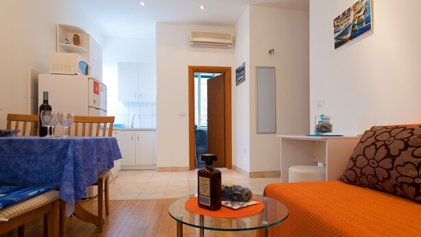 A modern living room with a glass coffee table, a sofa with an orange throw, and a dining area with a blue tablecloth. - Apartments Val (Split)