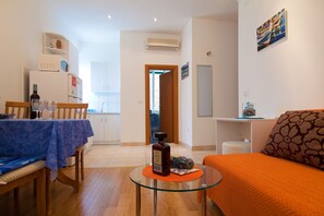 Una sala de estar moderna con una mesa de centro de vidrio, un sofá con un cojín naranja y un comedor con mantel azul.