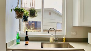A kitchen with white cabinets, a stainless steel dishwasher, a window with hanging plants, and a view of a building outside. - Birdhouse on GreenLake (Seattle)