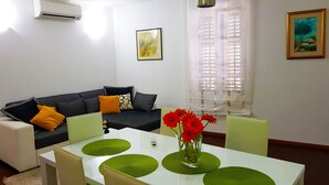 A modern living room with a grey sofa, a white dining table with green placemats, and a vase with red flowers.