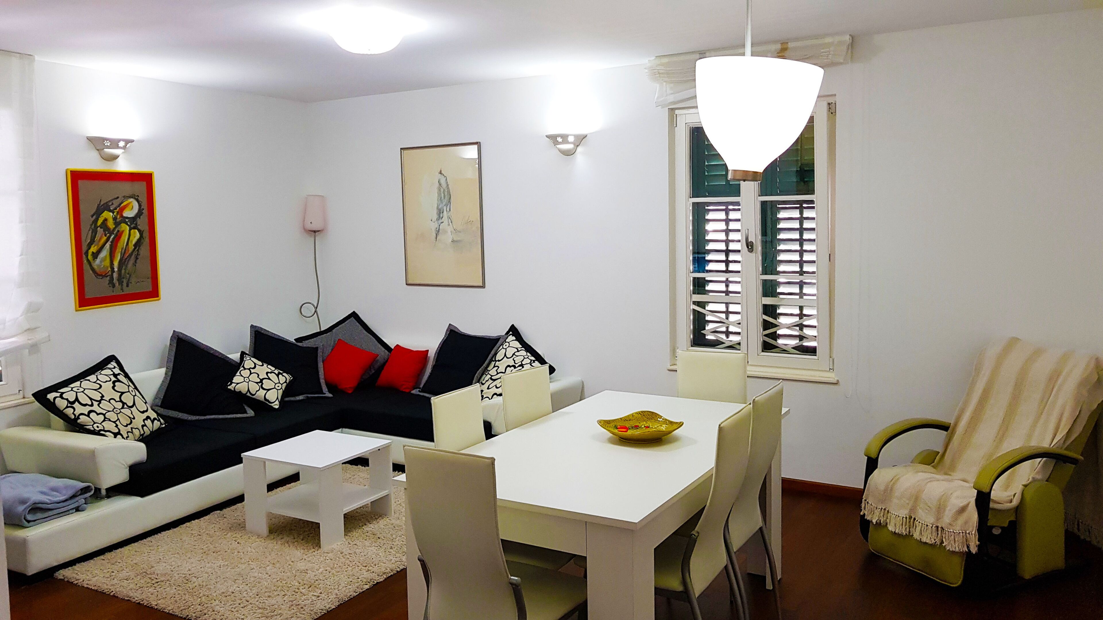 A living room with a sofa, a dining table, and a chair. There is a window with blinds, a painting on the wall, and a light fixture above the table.