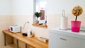 A kitchenette with a microwave, kettle, and a tea box. There is a wooden counter and a pink plant on the counter.
