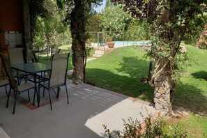 An outdoor patio with a table and chairs, a stone pillar, and a view of a swimming pool and garden.