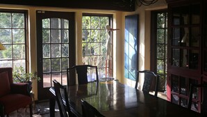 A dining area with a large table and chairs, large windows with views of greenery, and a cabinet with glass doors. - Historic English Country Lake House  (Lake Oswego)