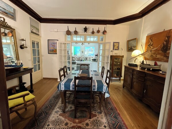 A dining area with a long table, chairs, and a patterned rug. There is a kitchen visible through an open door in the background.