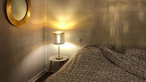 A bedroom with a patterned bedspread, a round mirror with a gold frame, a small bedside table with a lamp, and a hanging light fixture.