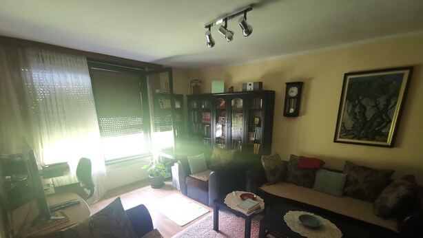 Ein Wohnzimmer mit Sofa, Sessel und Couchtisch. Ein Bücherregal, eine Uhr und ein gerahmtes Bild sind an der Wand zu sehen.