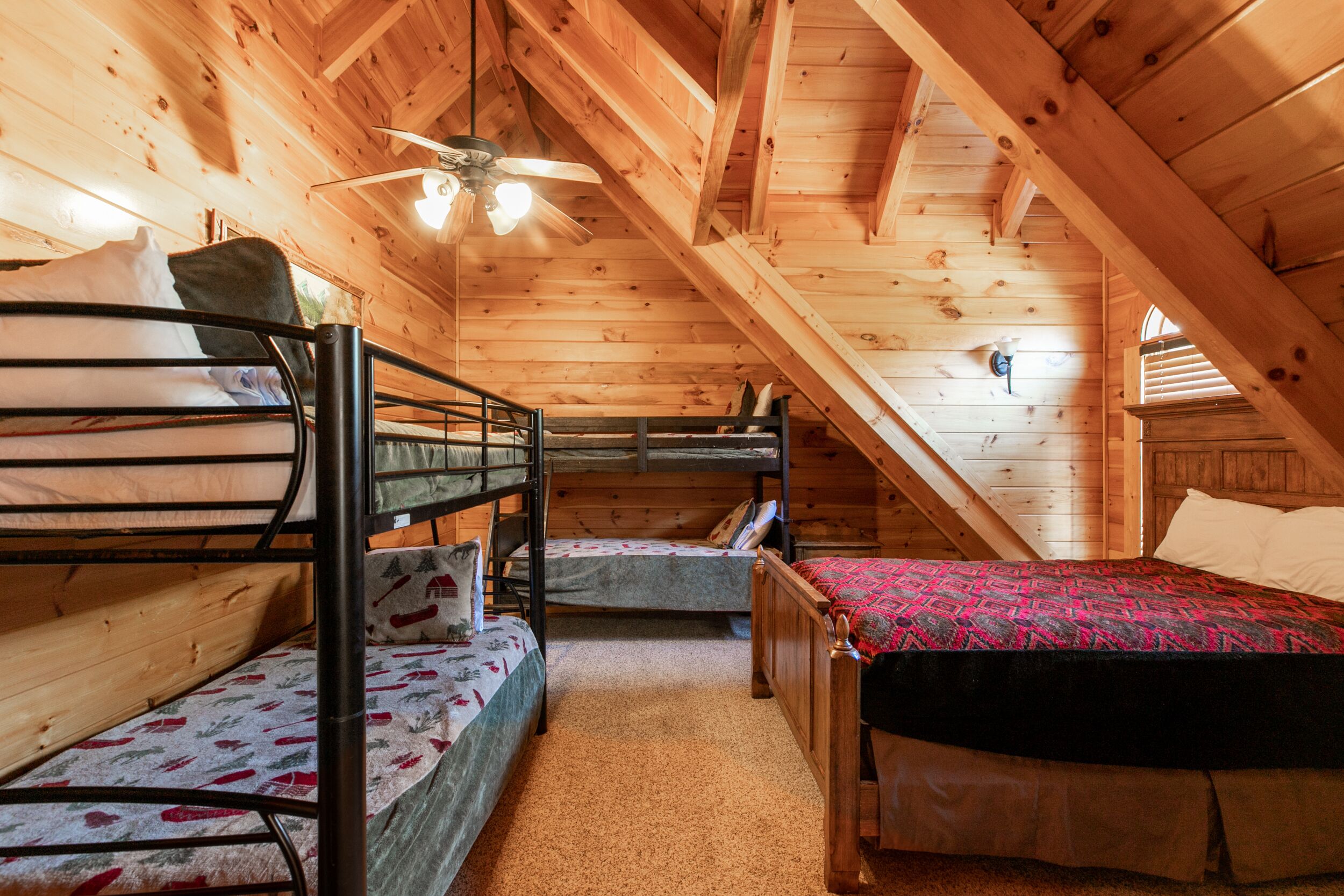 A cozy attic bedroom with wooden walls and a sloped ceiling. It features two bunk beds and a single bed, with a ceiling fan and a window providing natural light.