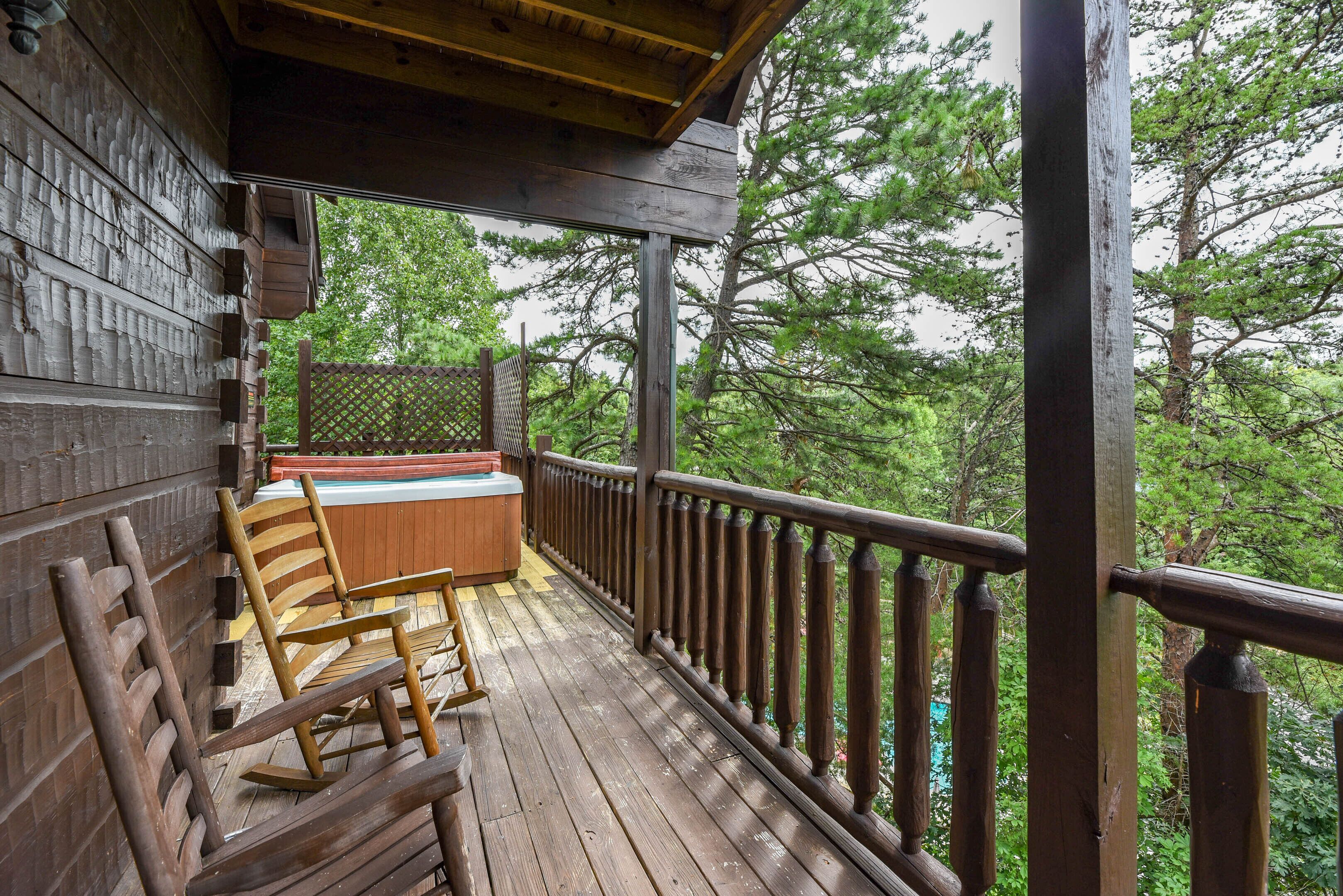 A wooden deck with a hot tub, wooden chairs, and a view of the forest.