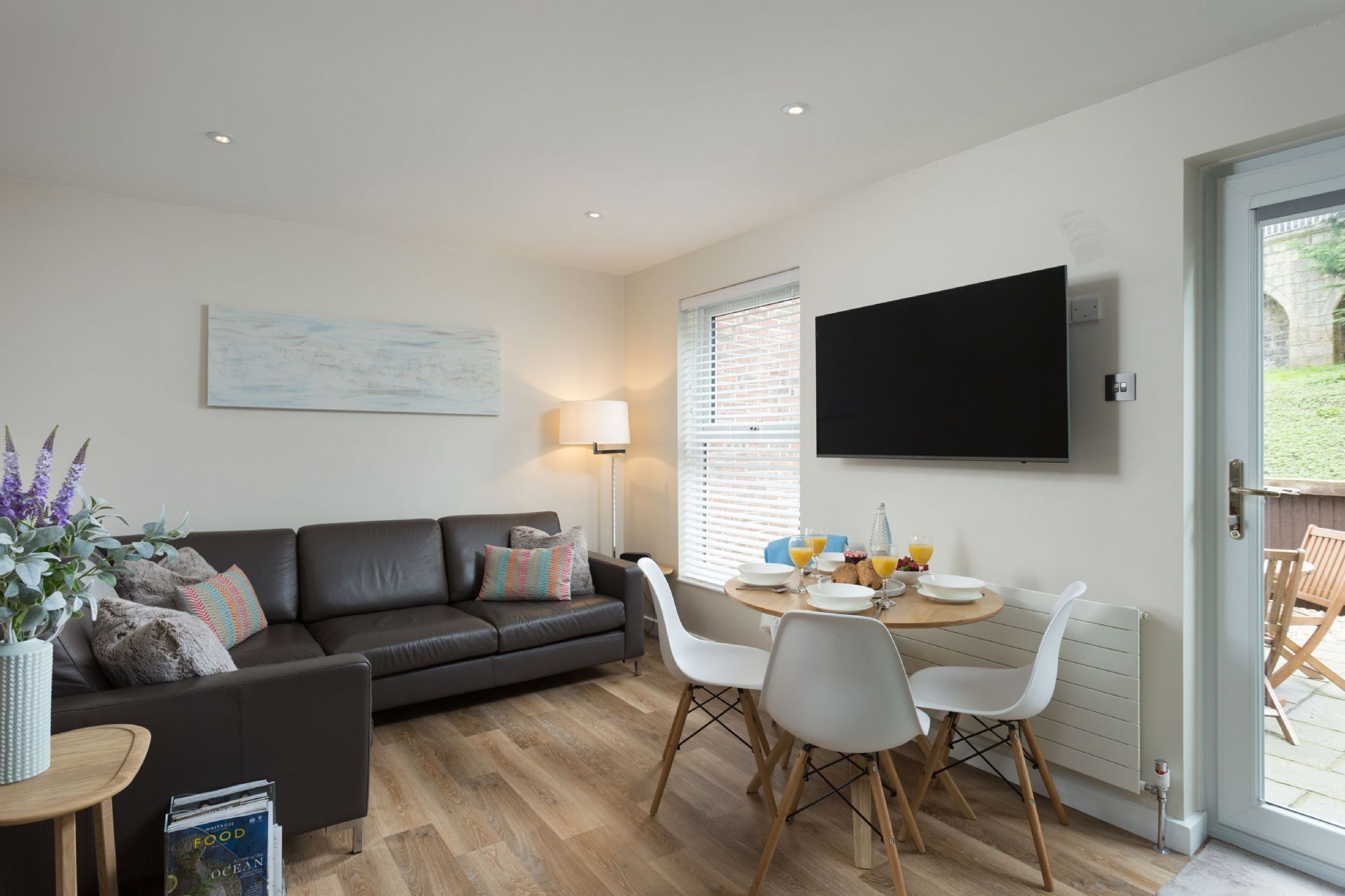 A modern living room with a dark leather sofa, a dining table with chairs, a flat-screen TV mounted on the wall, and a window with blinds.