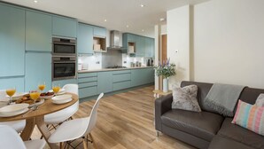 A modern kitchen with teal cabinets, a dining table with chairs, and a living area with a sofa and cushions. - 12 Pear Tree House - sleeps 4 guests  in 2 bedrooms (York)