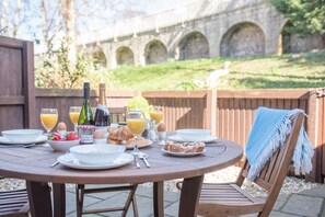 An outdoor dining table with breakfast items, a bottle of champagne, and a view of a stone bridge in the background. - 12 Pear Tree House - sleeps 4 guests  in 2 bedrooms (York)