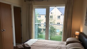 A bedroom with a large window, a bed with a beige comforter, two pillows, and a bedside lamp. - Penthouse Apartment on the River - Little Paxton (Saint Neots)