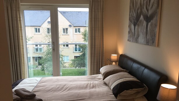 A hotel room with a bed, a nightstand, a lamp, and a view of a building through a window. - Penthouse Apartment on the River - Little Paxton (Saint Neots)
