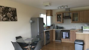 A modern kitchen with wooden cabinets, stainless steel appliances, and a dining area with a glass table and chairs. - Penthouse Apartment on the River - Little Paxton (Saint Neots)