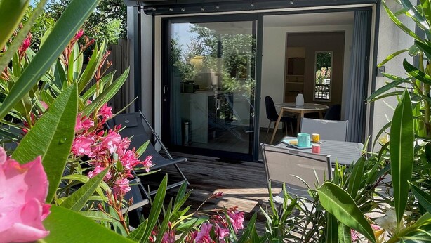 A modern outdoor dining area with a glass door leading to a room, surrounded by lush greenery and pink flowers. - Studio Anemone: self-catering cottage in Sarlat with private heated pool (Sarlat-la-Canéda)