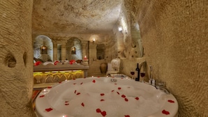 Una habitació de spa luxosa amb una banyera gran plena d'aigua i pètals de rosa, envoltada de parets de pedra i una decoració ornamentada.
