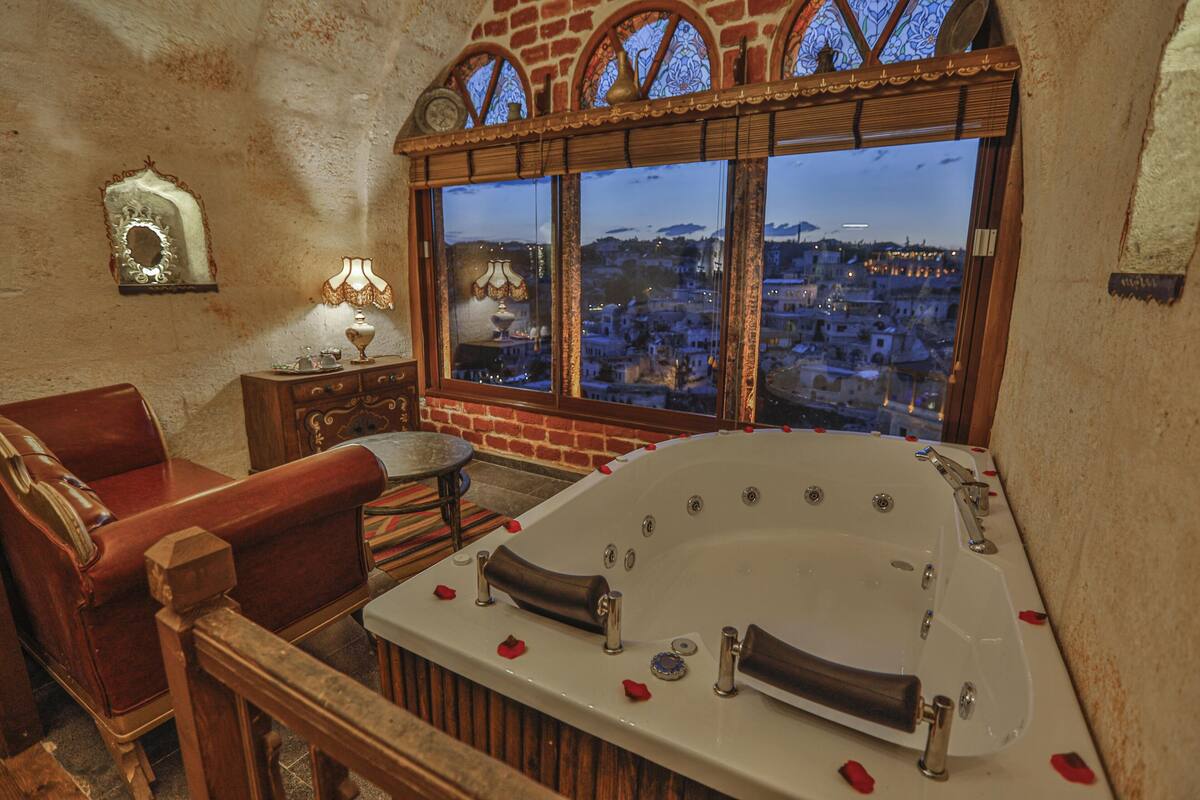 Ein Zimmer mit einer großen Badewanne, einem Holzstuhl, einem kleinen Tisch mit einer Lampe und Blick auf eine Stadtlandschaft durch Bogenfenster.