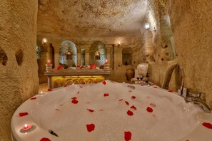 Un spa lujoso con una bañera grande llena de agua y pétalos de rosa, un espejo y una mesa con copas de vino.