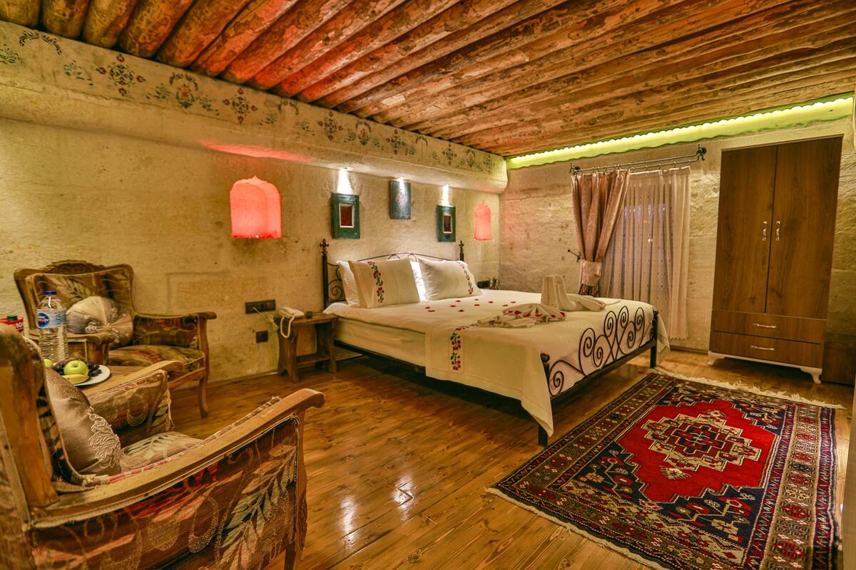 Ein traditionell eingerichteter Raum mit Holzboden, einem Bett mit floralem Überwurf, einem Holzstuhl, einem Nachttisch und einem roten Teppich mit Muster.