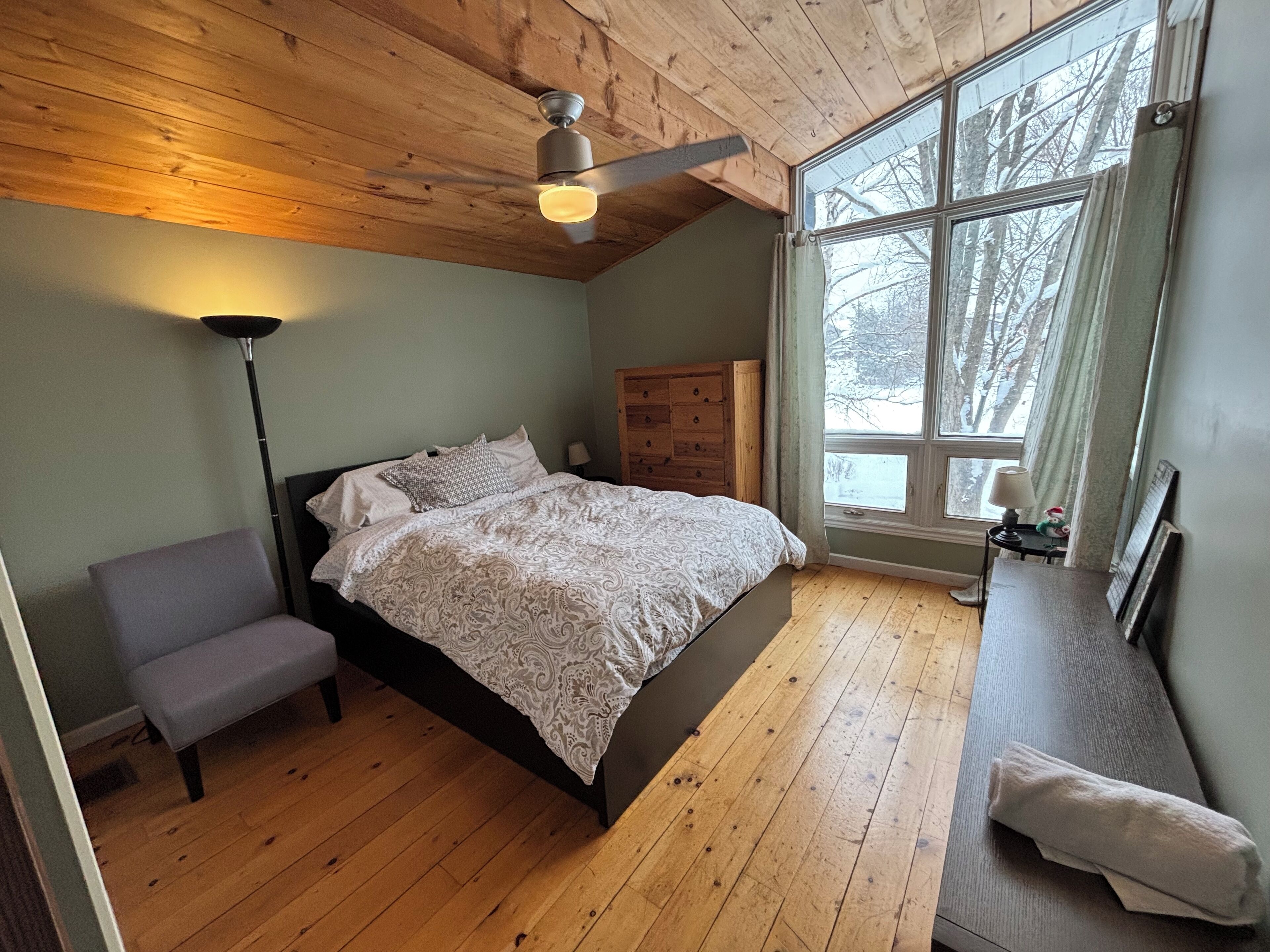 Une chambre avec un plafond en bois, un grand lit avec une literie à motifs, une table de chevet, une lampe et une fenêtre donnant sur des arbres.