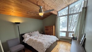 Ein Schlafzimmer mit Holzbalkendecke, einem großen Bett mit gemusterten Betttextilien, einem Nachttisch, einer Lampe und einem Fenster mit Blick auf Bäume.