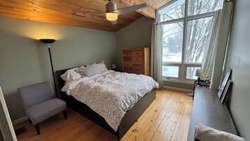 Une chambre avec un plafond en bois, un grand lit avec une literie à motifs, une table de chevet, une lampe et une fenêtre donnant sur des arbres.
