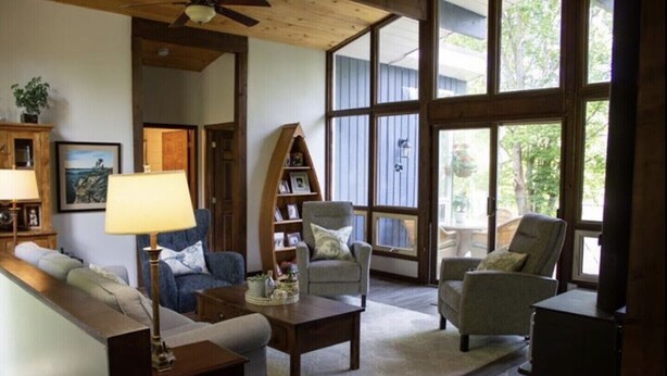 Ein gemütliches Wohnzimmer mit einer Couch, Sesseln, einem Couchtisch und einem Bücherregal. Große Fenster lassen natürliches Licht herein und bieten Ausblicke ins Freie.